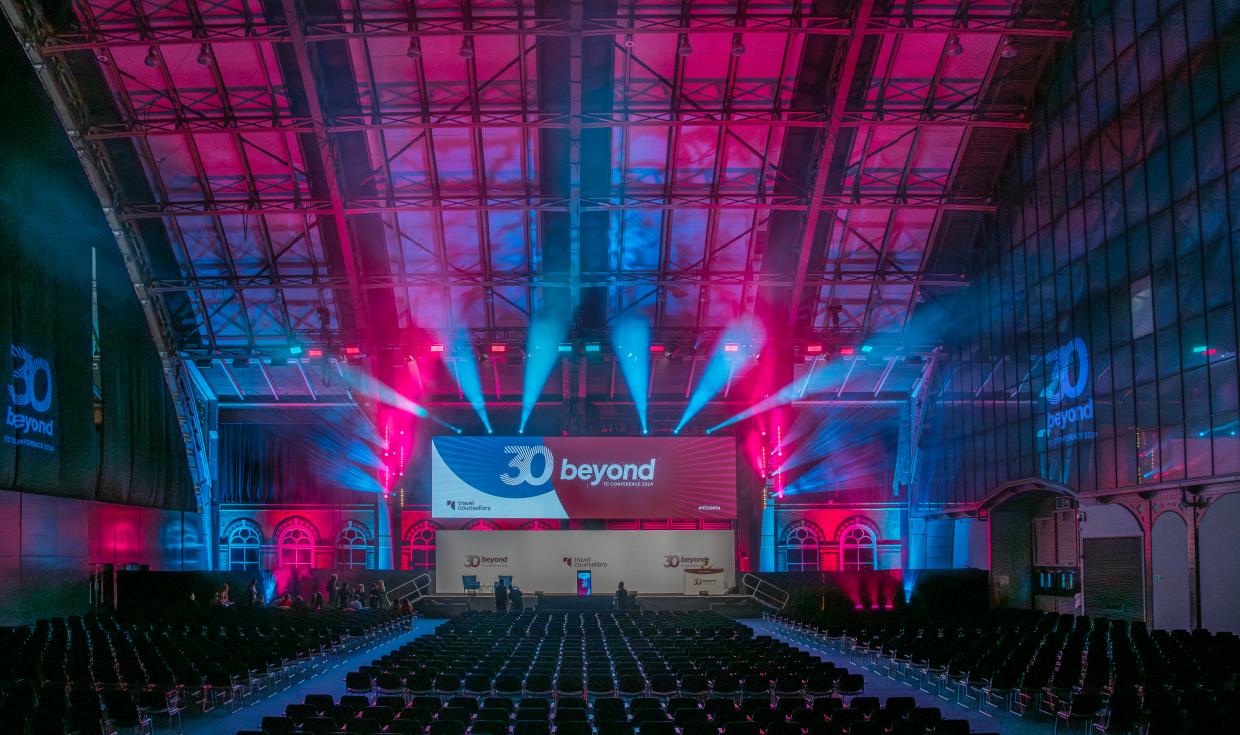 Large stage lit up with pink and blue lighting with lines of seats facing it, with a screen reading 30 beyond