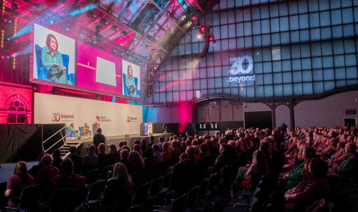 A large audience watching a speaker panel on stage, lit up with pink and blue lights, under the arched ceiling of Manchester Central.