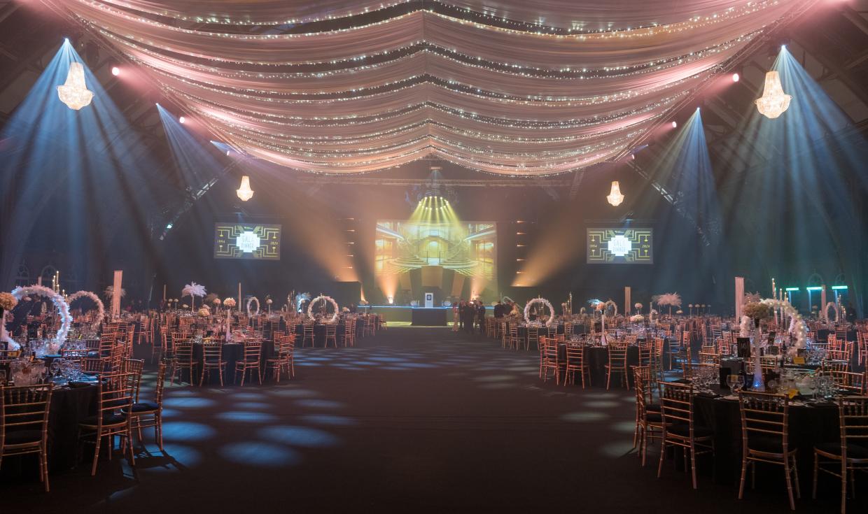 Round tables set up for a gala dinner, all lit up from the ceiling, with a stage at the end of the room.