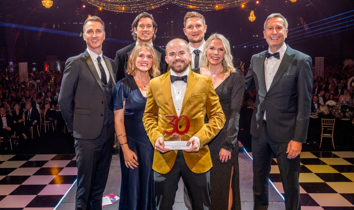 Individuals stood together on stage with an award, with attendees in the background at tables behind them..jpg